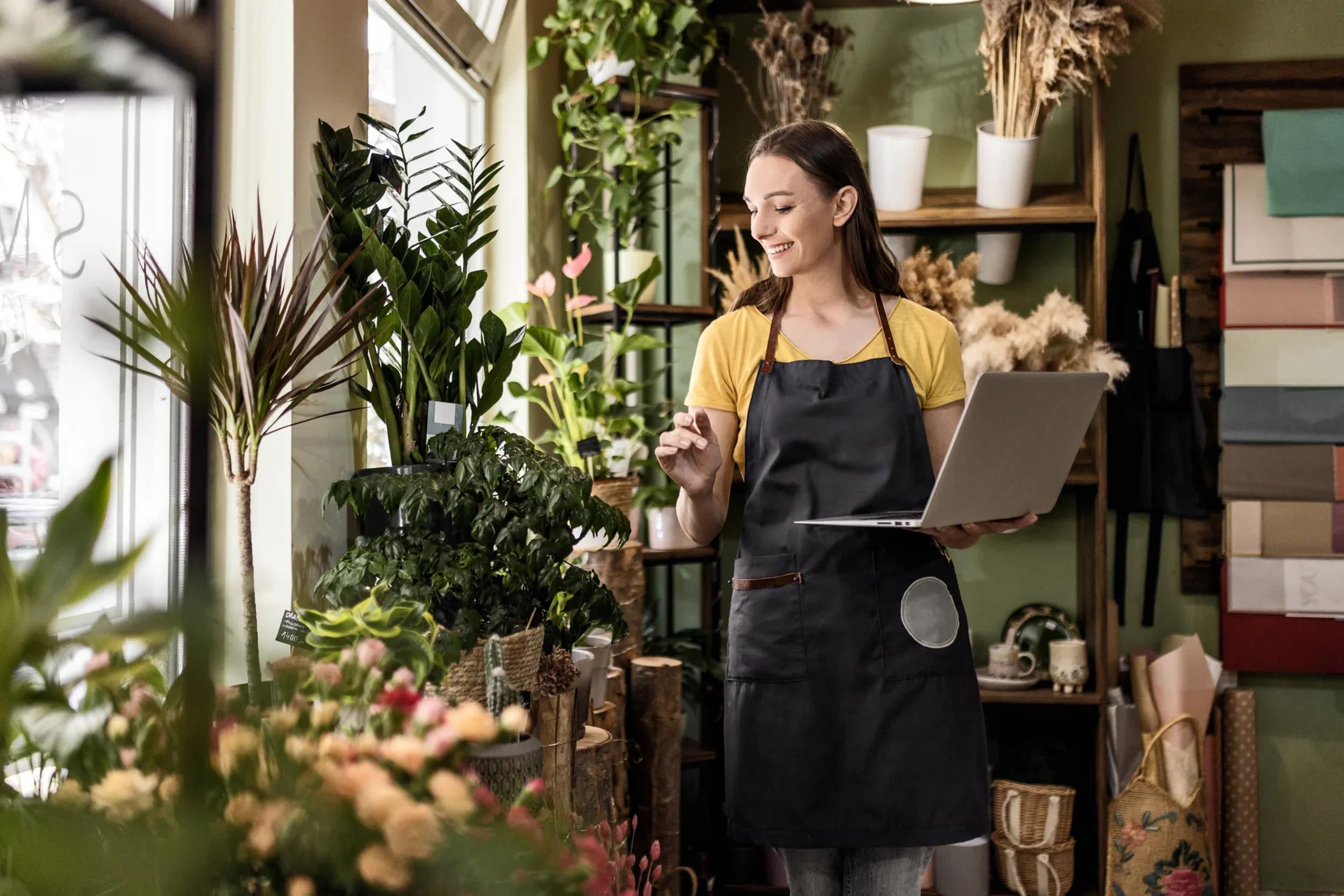 young woman is standing among plants and flowers in her florist shop. she is smiling and holding a laptop in her left arm while tending to a plant with her other arm. she is wearing a pale yellow top and black apron.