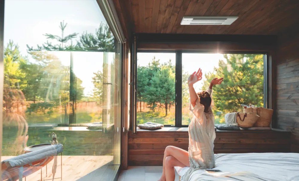 woman sitting on bed in the morning looking out her large windows at a green nature setting with trees
