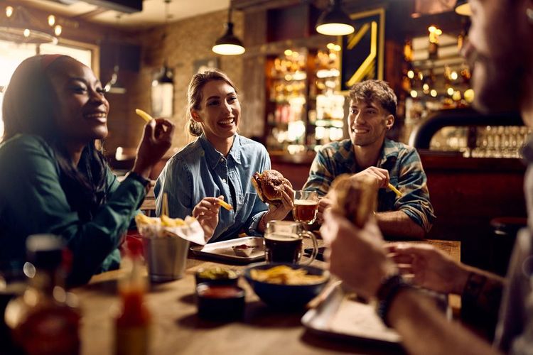 group of young people are sitting around a table in a cozy restaurant. they are enjoying hamburgers and french fries.